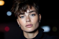 Close up portrait of young androgynous model with striking blue eyes and short dark hair against blurred night lights background, showcasing natural makeup and pierced ears.
