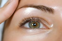 Close-up macro shot of green eye with long natural eyelashes and well-groomed eyebrow. Fingers gently touching smooth skin around eye area. Clean beauty concept.