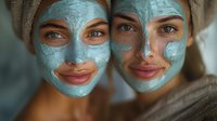 Two young multicultural women wearing blue clay face masks and hair towels smile during spa skincare treatment. Natural beauty and wellness concept for cosmetic advertising.