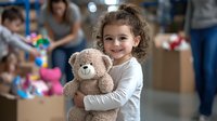 Cheerful young girl with curly hair hugging plush teddy bear in playroom setting, wearing white long sleeve shirt. Natural candid childhood moment.