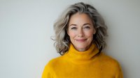 Confident mature woman with gray hair wearing yellow turtleneck sweater smiling at camera against light background, natural beauty and aging gracefully concept.