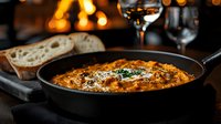 Creamy tomato risotto with herbs served in cast iron skillet alongside crusty bread and wine glass, cozy fireplace dining atmosphere for comfort food lovers.