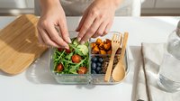 Woman's hands arranging fresh green salad with tomatoes and berries in glass containers on wooden tray for meal prep and healthy eating.