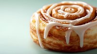 Fresh baked cinnamon roll with white vanilla glaze dripping down spiral layers, close-up view on mint green background showing soft texture and gooey topping.