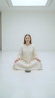 Young woman of Caucasian ethnicity meditating in lotus position on white floor in minimalist interior space with skylight, wearing beige linen dress, practicing mindfulness and yoga.
