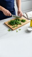 Male chef chopping fresh green herbs on wooden cutting board in modern kitchen, with olive oil and salt nearby.