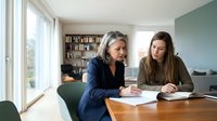 Professional mature woman mentoring young female colleague at modern office desk reviewing documents together in bright workspace with natural lighting.