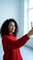 Happy young African American woman in red sweater taking selfie with smartphone near window in bright modern interior for social media content.