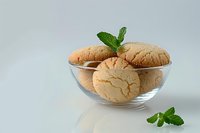 Fresh baked sugar cookies in clear glass bowl garnished with mint leaves on light background, minimalist food photography composition.