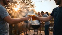 Friends toasting with beer glasses at outdoor barbecue party during golden hour sunset with string lights creating warm festive atmosphere for celebration.