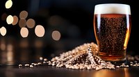 Glass of fresh craft beer with bubbles and foam head next to scattered barley grains and wheat ears on dark wooden surface with blurred bokeh lights background.