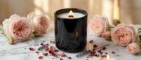 Luxury black candle burning with soft flame surrounded by pink roses and scattered petals on marble surface creating romantic spa atmosphere for relaxation.