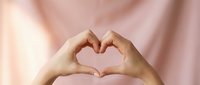 Hands forming a heart shape gesture against a soft pink background, symbolizing love, care, and support for health and wellness campaigns.