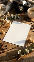 Blank white paper card on rustic wooden table with vintage camera, coffee cup, dried flowers and eucalyptus leaves in warm natural sunlight for mockup design.