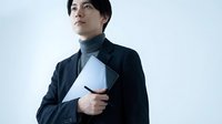 Young East Asian man in dark navy blazer holding white laptop, looking upward thoughtfully against neutral background. Suitable for business, technology, and professional concepts.