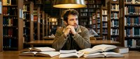 Young Caucasian male student studying in university library surrounded by open books and bookshelves, academic research and education concept.