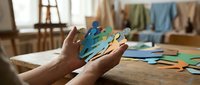 Hands holding colorful paper puzzle pieces in bright workshop setting with crafting materials and creative workspace for teamwork concepts.