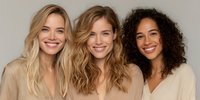 Three diverse women with natural makeup smiling together in beige clothing against neutral background for lifestyle and beauty campaigns.