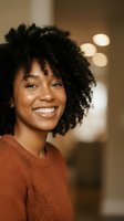 Happy young African American woman with curly hair smiling warmly in cozy indoor setting with soft lighting for lifestyle and wellness content.