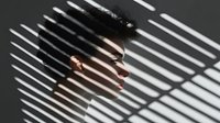 Young Asian woman's face partially hidden behind window blinds casting dramatic shadow lines, creating mysterious noir atmosphere and visual contrast.