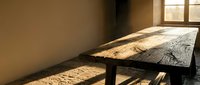 Wooden table with natural live edge top positioned near sunlit window, casting long shadows across floor in minimalist interior space.