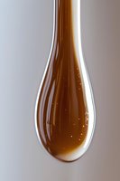 Macro shot of golden honey or syrup drop forming teardrop shape with tiny air bubbles against light background, showing viscous liquid texture and natural amber color.