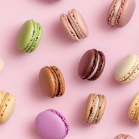 Colorful French macarons scattered on pink background in minimal pattern layout, featuring pastel and chocolate shades for confectionery and bakery promotion.