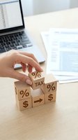 Hand placing wooden cube with percentage symbol on stack of tax and financial blocks on office desk with laptop and documents for accounting concepts.