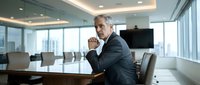 Mature male executive in dark suit sitting at conference table in modern office boardroom with floor-to-ceiling windows overlooking cityscape, suitable for corporate business concepts.