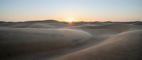 Golden sunrise over vast sand dunes creating dramatic shadows and warm light across desert landscape with rolling hills in background for travel content.