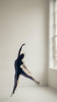 Female ballet dancer in black leotard performing grand jeté leap in minimalist white studio space with natural window light, capturing graceful movement and athletic form.