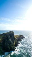 Dramatic coastal cliffs with basalt rock formations rise above turquoise ocean waters under clear blue sky. Lighthouse stands on distant headland. Ideal for travel, nature, and scenic backgrounds.