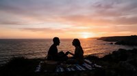 Silhouetted couple sitting on rocky coastline at sunset, watching golden light reflect on calm ocean waters during romantic evening by the sea.