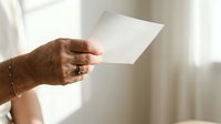 Close-up of mature woman's hand holding blank white card or letter against bright window light, suitable for greeting, invitation, or business communication concepts.