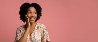 Young African American woman with curly hair smiling while talking on smartphone against pink background, expressing joy and positive emotions during phone call.