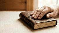 Elderly hands resting on worn leather-bound book in warm sunlight, symbolizing wisdom, knowledge, faith, and life experience in peaceful moment.