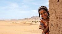 Young girl with dark hair and medium brown skin tone, wearing colorful traditional clothing, peeking around adobe wall in desert landscape with distant mountains and settlements.