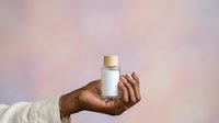 Cosmetic product mockup with hand holding white bottle and gold cap against soft pink blurred background for skincare branding and packaging design.