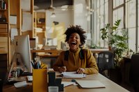 Young African American woman with natural curly hair laughing while working at desk in modern coworking space, wearing mustard sweater, expressing joy and happiness.