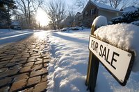 Snow covered residential street with for sale sign in winter sunlight, cobblestone driveway leading to suburban house with dramatic morning sun rays creating long shadows.