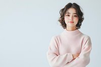 Young Asian woman with short wavy hair wearing soft pink sweater standing with crossed arms against light background, expressing confidence and gentle smile.
