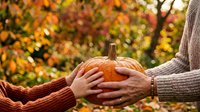 Couple holding large orange pumpkin together outdoors in autumn garden with blurred fall foliage background, seasonal harvest celebration and togetherness concept for holiday marketing.