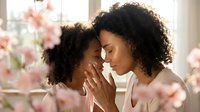 Mother and daughter with curly hair bonding indoors near pink flowers, showing affection and togetherness for family, parenting, and lifestyle content.
