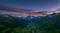 Dramatic alpine valley landscape at sunset with snow-capped mountain peaks, lush green slopes, and small villages nestled in the terrain below, suitable for travel and nature publications.