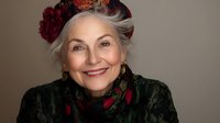Elegant mature woman with silver hair wearing floral crown and dark patterned dress smiles warmly at camera against neutral background. Portrait captures natural aging beauty.