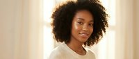 Young African American woman with curly hair smiling warmly in cozy cream sweater by window with soft natural lighting for lifestyle and portrait photography.