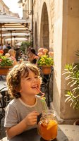 Happy curly-haired child enjoying fresh orange juice at outdoor Mediterranean cafe terrace with stone architecture and warm sunlight creating joyful summer moment.