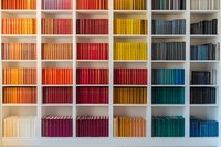 Colorful books arranged on white shelves creating rainbow gradient pattern from orange to blue. Modern library or bookstore interior design showcases organized collection.