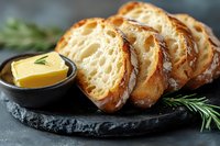 Fresh crusty bread slices with butter and rosemary served on black stone plate, close up view of artisanal baked goods with golden crust and airy texture.