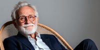 Senior Caucasian man with white beard and glasses relaxing in wicker chair, wearing navy blazer and white shirt, expressing warmth and wisdom against neutral background.